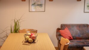 A dining table with a wire fruit basket, a wooden chair, and a sofa with a red pillow in a room with wooden beams and a framed picture on the wall.
