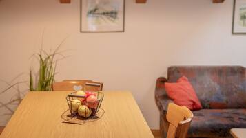 A dining table with a wire fruit basket, a wooden chair, and a sofa with a red pillow in a room with wooden beams and a framed picture on the wall.