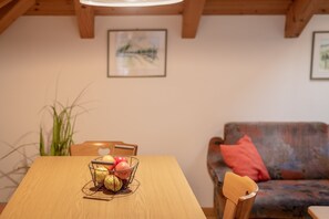 A dining table with a wire fruit basket, a wooden chair, and a sofa with a red pillow in a room with wooden beams and a framed picture on the wall.