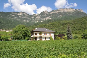 Una casa de dos pisos con un balcón, rodeada por un viñedo, con montañas al fondo.