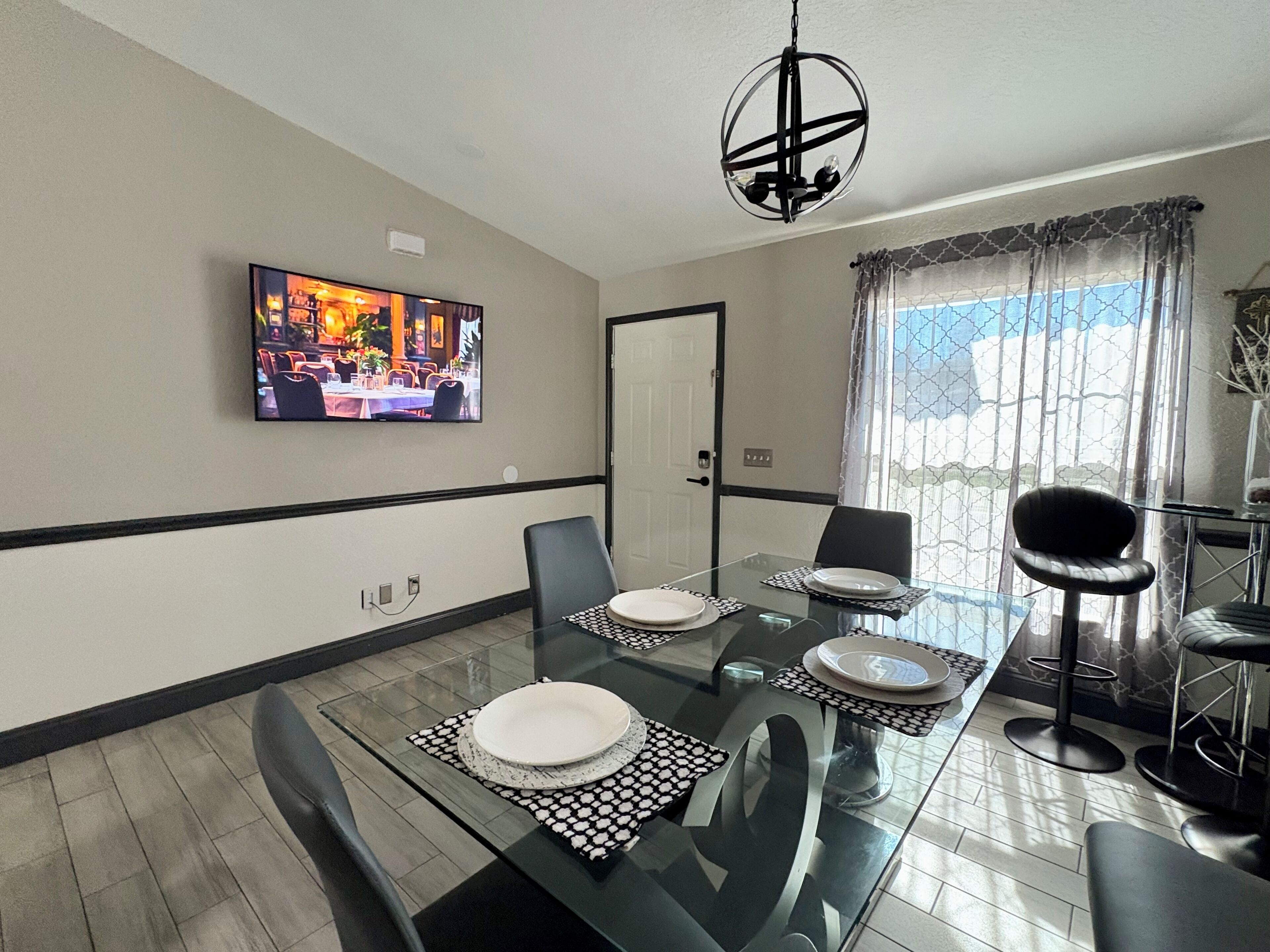 A dining area with a glass table, black and white patterned placemats, and black chairs. A television is mounted on the wall displaying a restaurant scene.