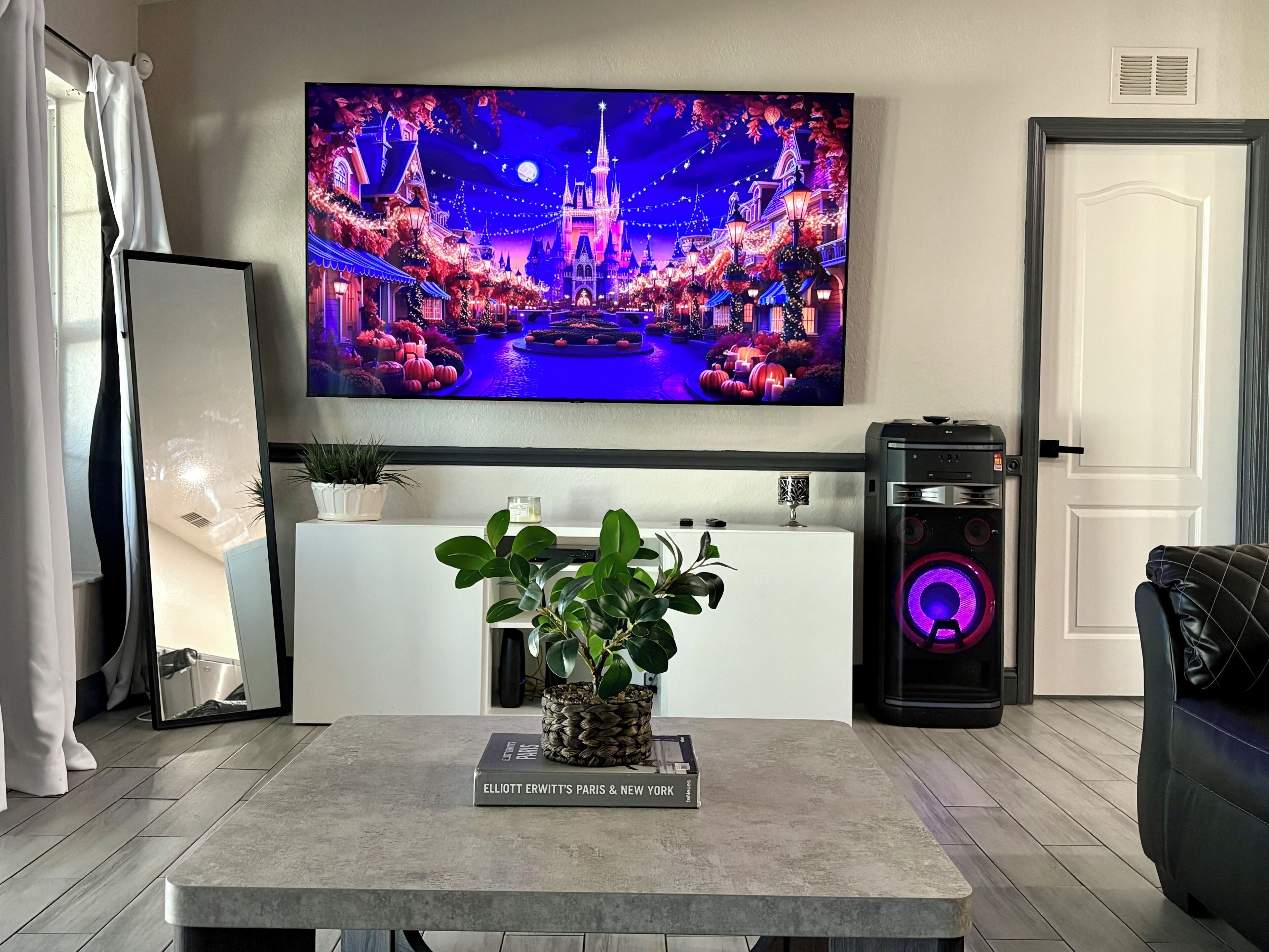 A living room with a flat-screen TV displaying a festive scene, a coffee table with a book, a plant, and a speaker.