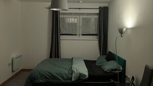 Une petite pièce faiblement éclairée, avec un lit simple, une chaise, un bureau et une fenêtre avec des rideaux.