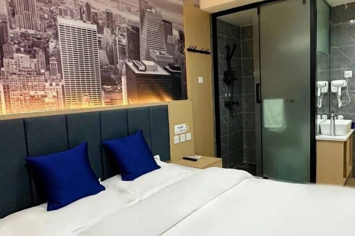 Hotelkamer met een bed, twee blauwe kussens, een stadsgezicht op de muur, een glazen douche en een nachtkastje met een telefoon.