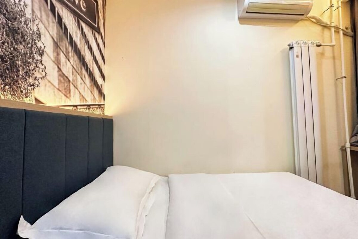 A neatly made bed with white linens in a room with a plain wall and an air conditioning unit above it.