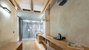 Una camera d'albergo moderna con una panca in legno, una scrivania con bollitore e tazze, e un box doccia in vetro.