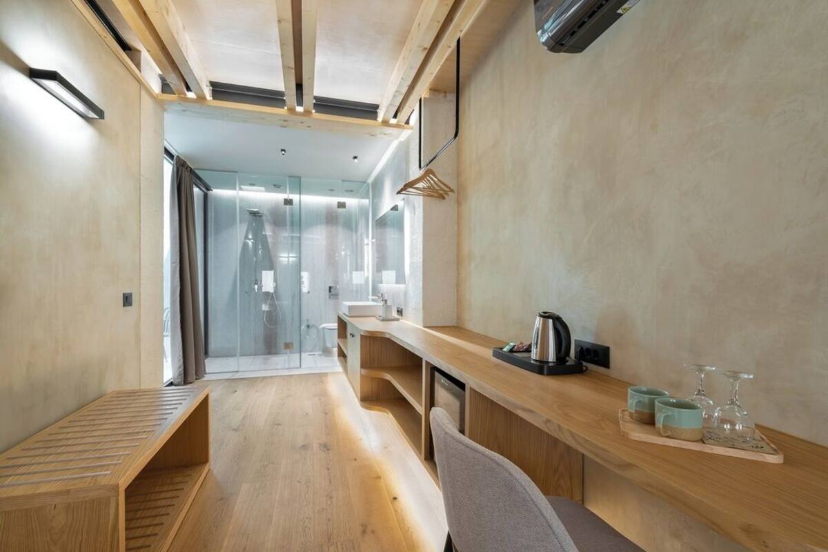 A modern hotel room with a wooden bench, a desk with a kettle and mugs, and a glass shower enclosure.
