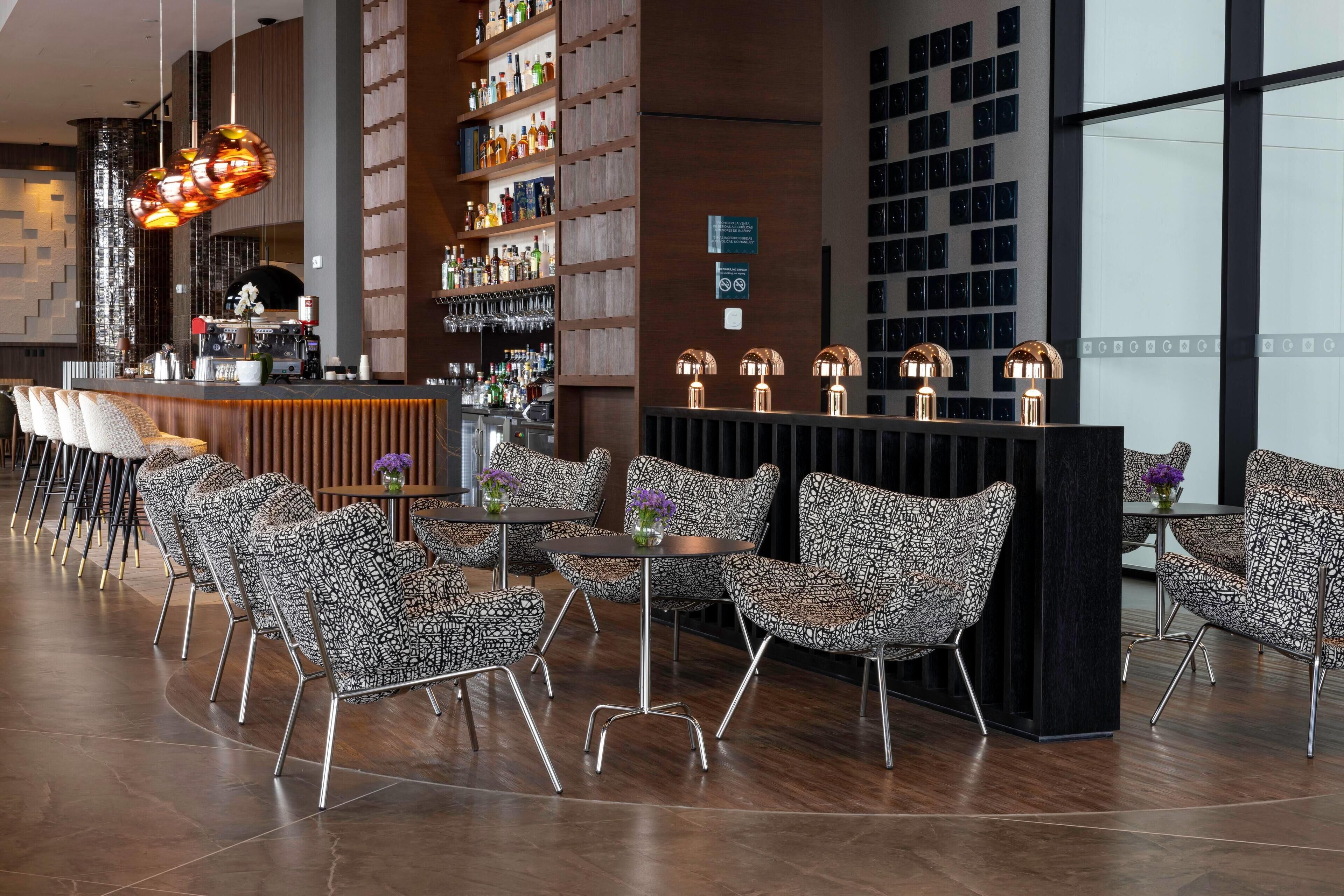 A modern bar area with patterned chairs, a counter with a coffee machine, and shelves stocked with bottles.