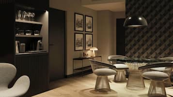 A modern interior with a dining area featuring wireframe chairs and a glass-top table, a bookshelf with decorative items, and framed cityscape pictures on the wall.