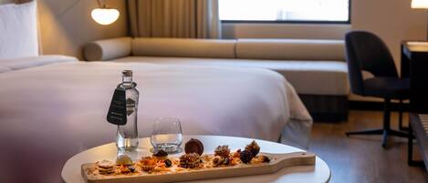A hotel room with a neatly made bed, a bedside table with a bottle and glass, and a tray of assorted pastries.