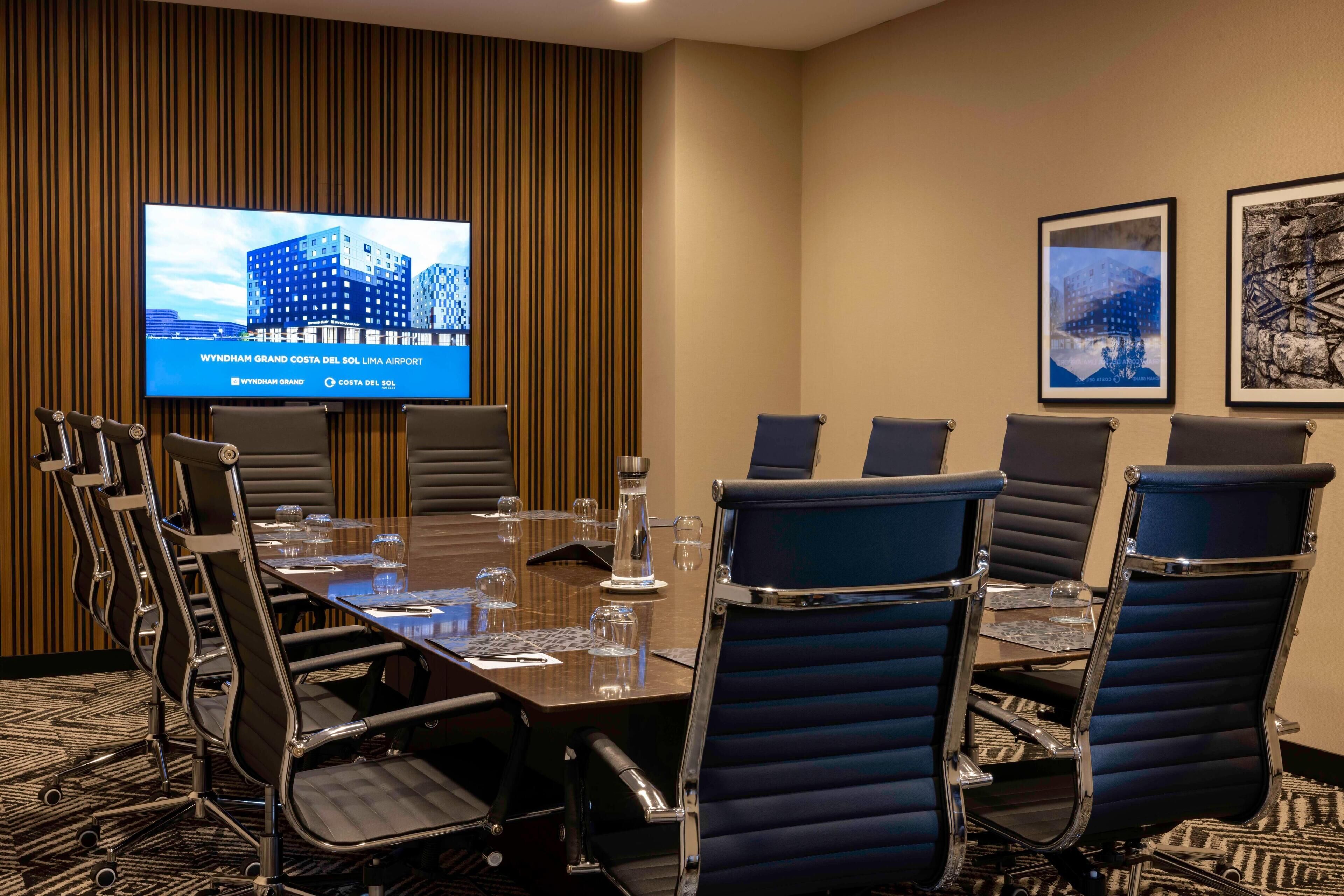 Un salón de conferencias con una mesa larga, seis sillas, una pantalla de televisión y cuadros en la pared.