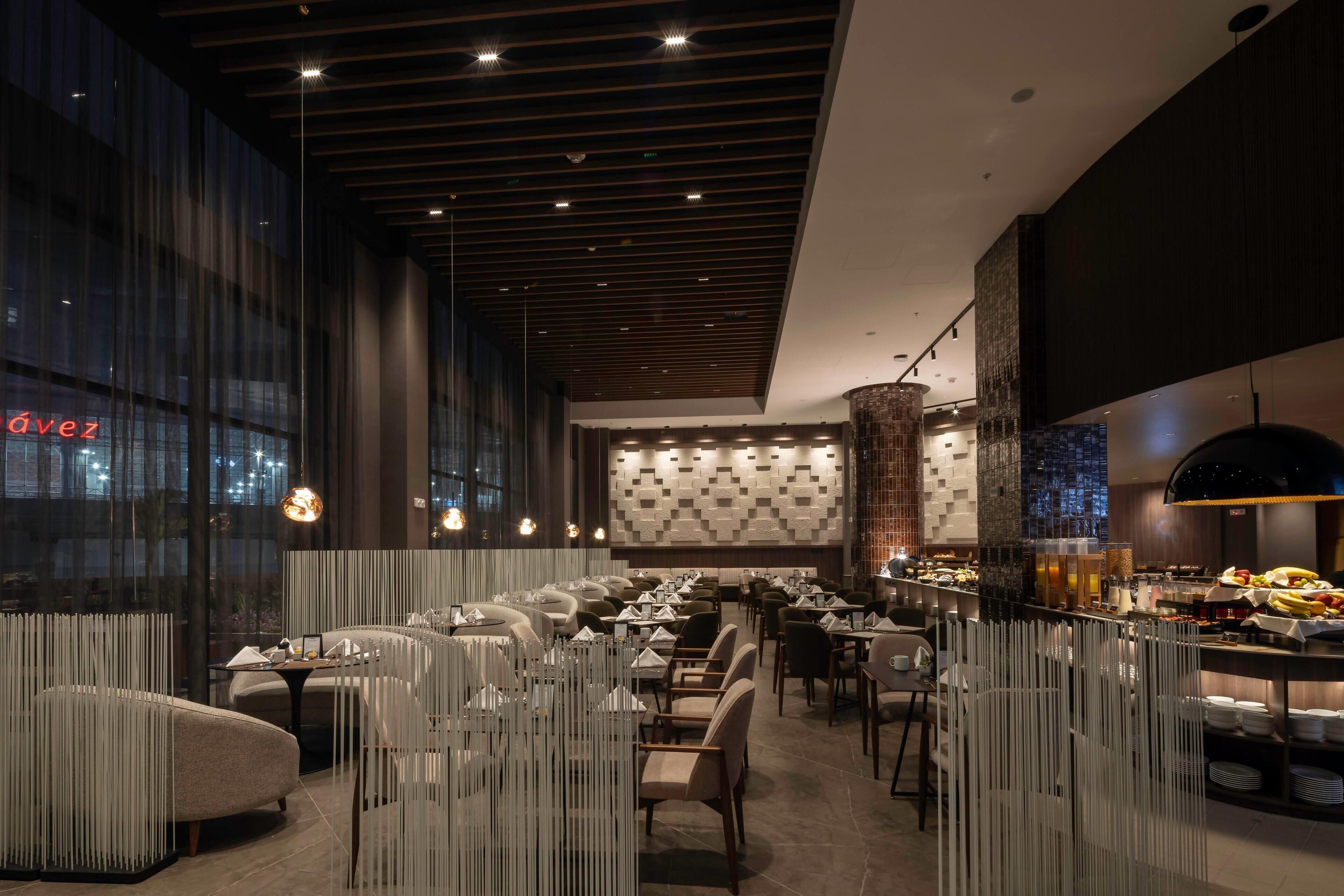 Een modern restaurant met een lange eetzône, hoge plafonds en een patroonwand op de achtergrond.
