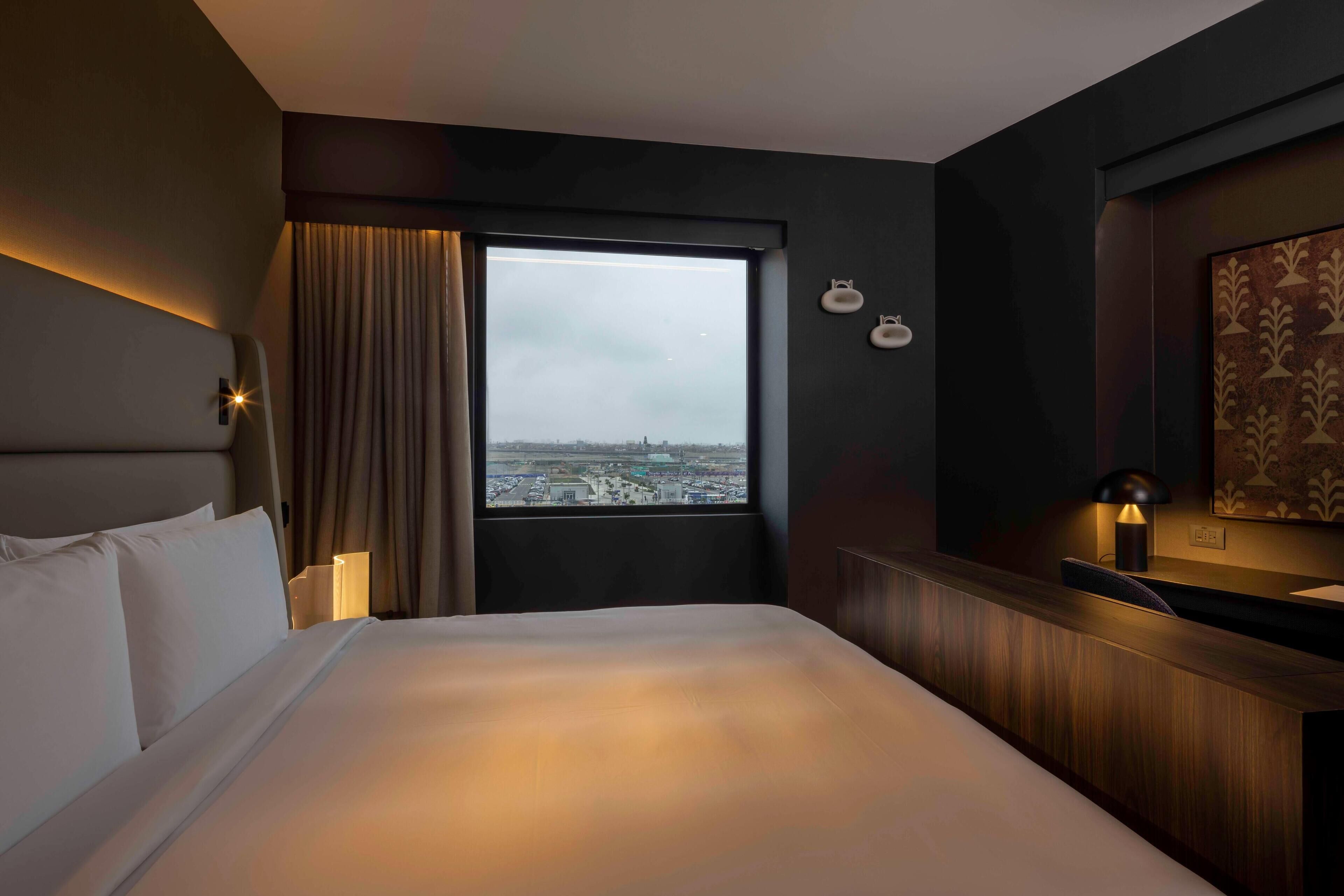 A hotel room with a large bed, a nightstand, a lamp, a window with a view of the city, and a wall-mounted light fixture.