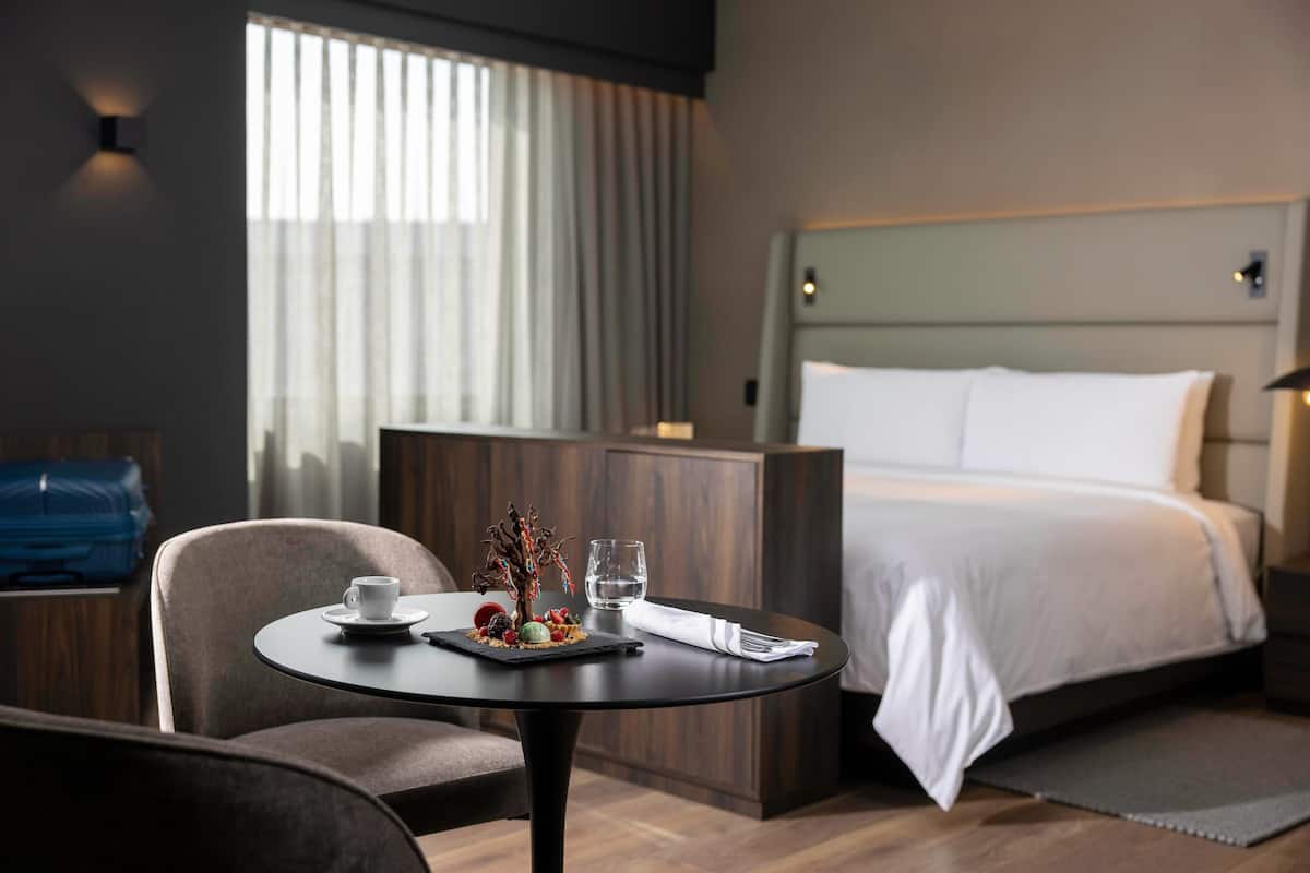Une chambre d’hôtel avec un lit, une table ronde sur laquelle se trouvent une tasse, un verre et une corbeille de fruits.