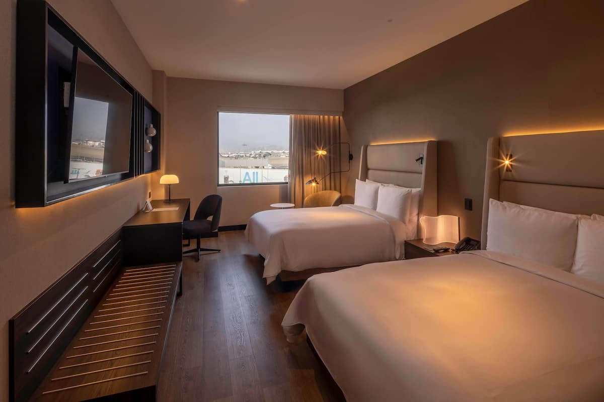 Une chambre d’hôtel avec deux lits, un bureau, une chaise, une grande fenêtre et une vue sur la ville.