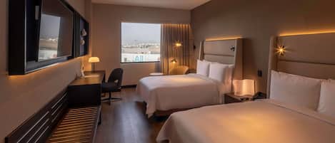 A hotel room with two beds, a desk, a chair, a large window, and a view of the city.