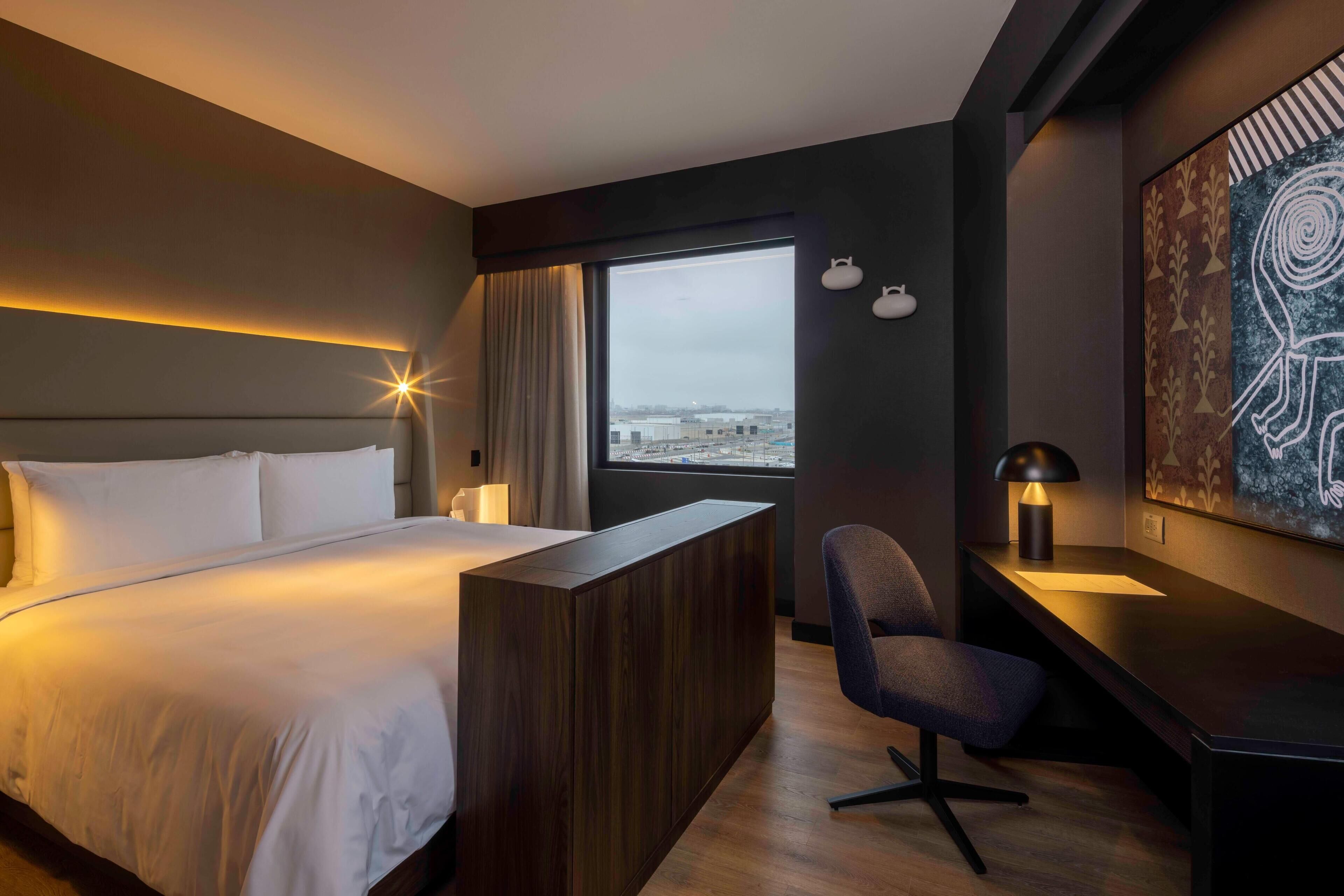 A modern hotel room with a large bed, a desk, and a chair. There is a window with a view of the city.