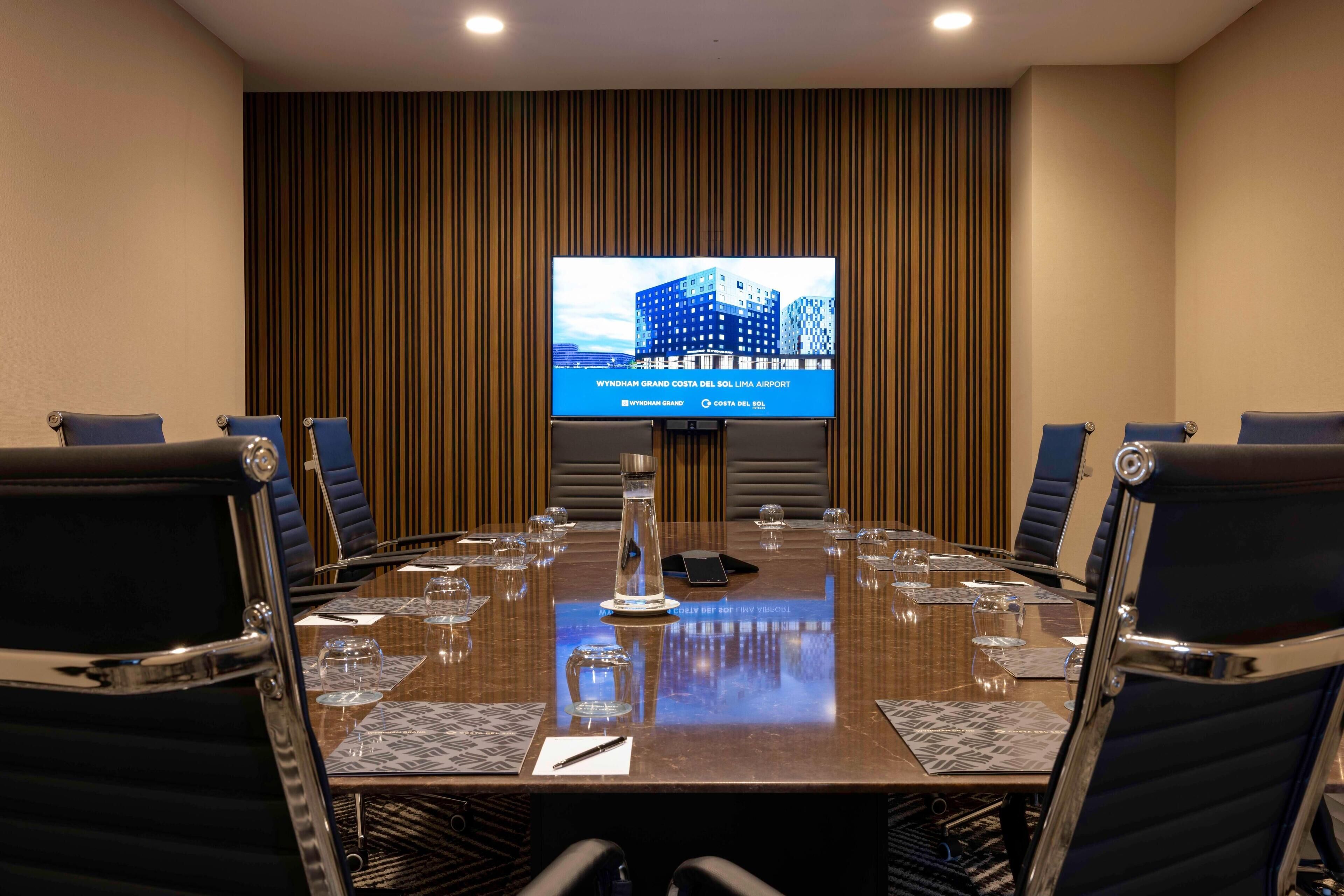 Un salón de conferencias con una mesa grande, sillas, una pantalla que muestra una imagen de un aeropuerto y una botella de agua.