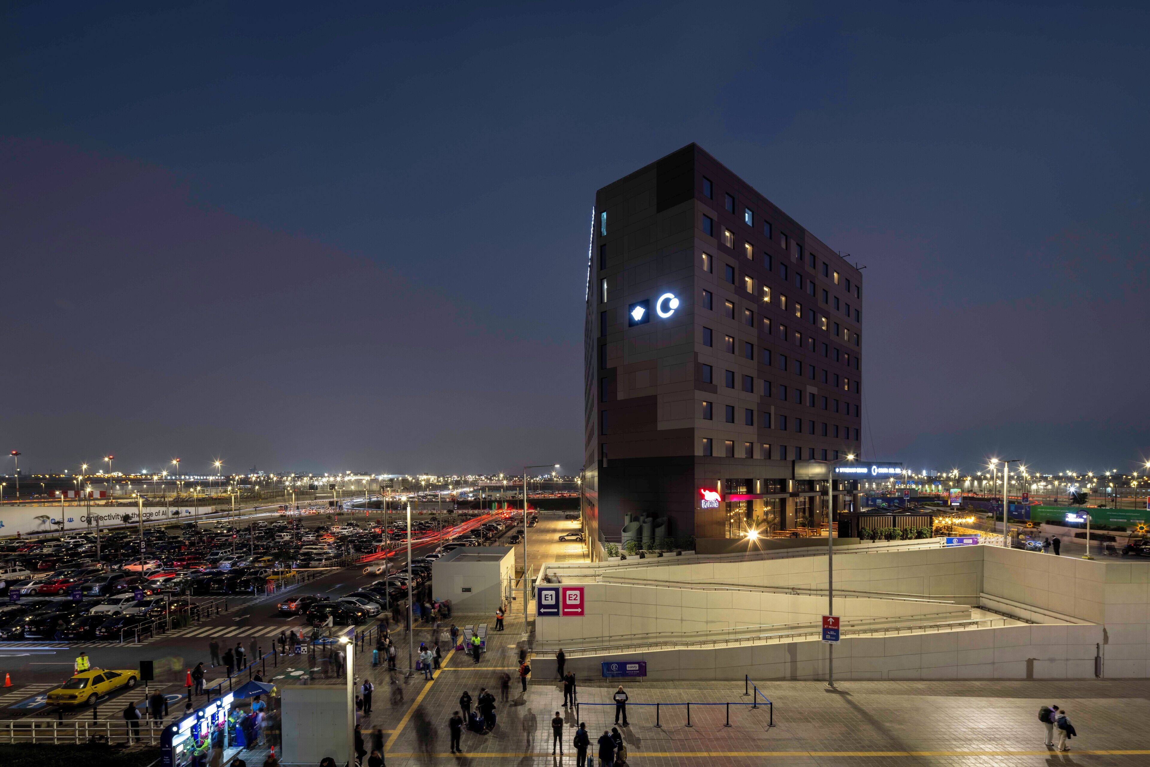 A modern hotel with a unique angular design, located near a busy airport with a parking lot and terminal in the background.