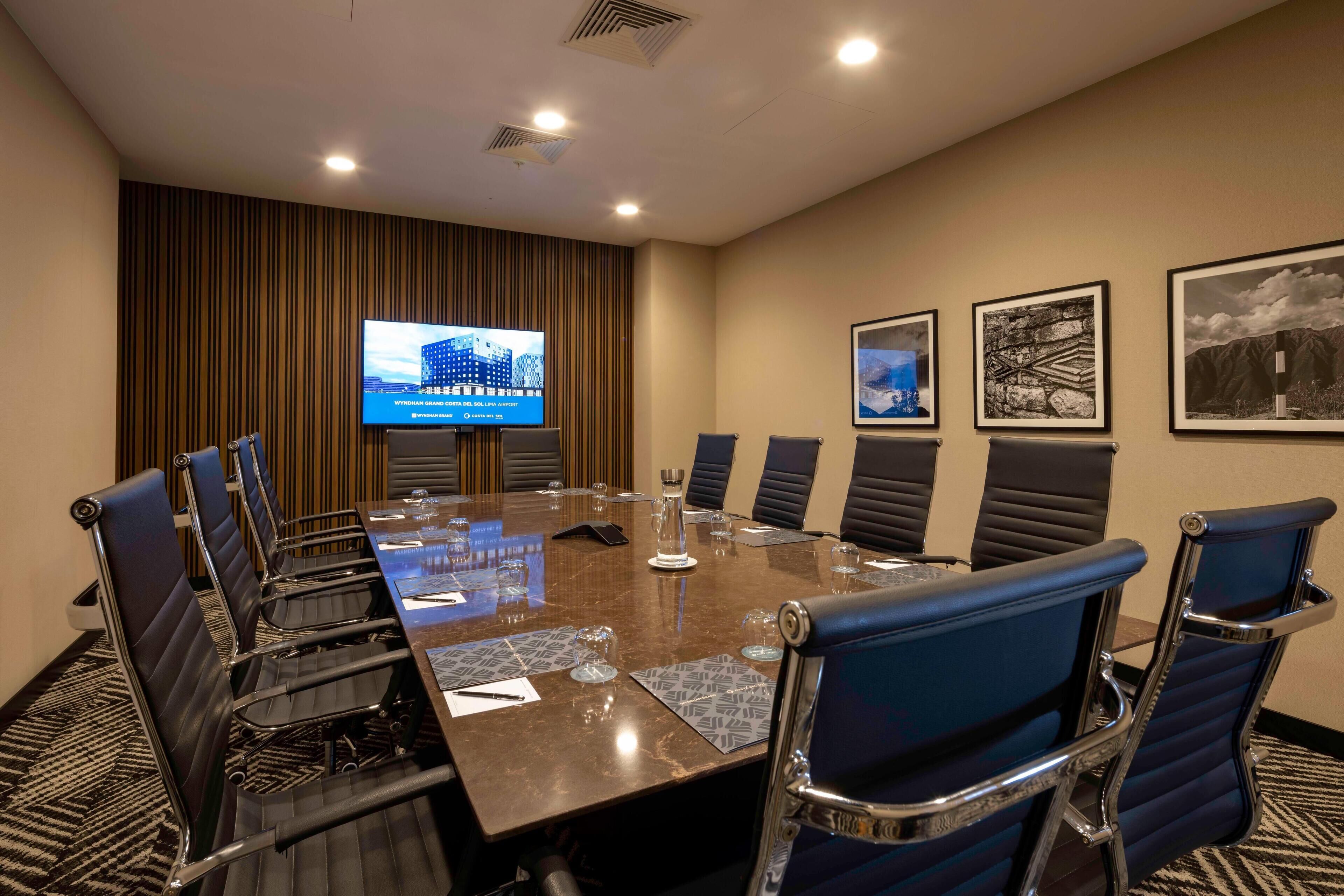 Un salón de conferencias con una gran mesa, sillas, una pantalla de televisión y cuadros en la pared.