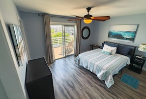 A bedroom with a bed, a ceiling fan, a view of the outdoors, and a painting on the wall.