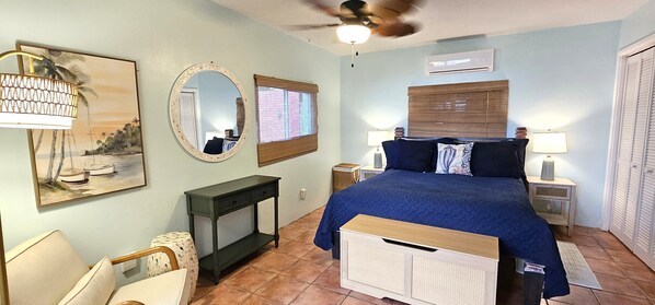 Een slaapkamer met een bed, een bureau, een stoel, een spiegel, een raam met jaloezieën en een plafondventilator.