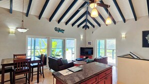 Una cocina amplia con una isla grande, comedor con sillas y sala de estar con sofá. Hay ventanas amplias que ofrecen luz natural y vistas a la vegetación.