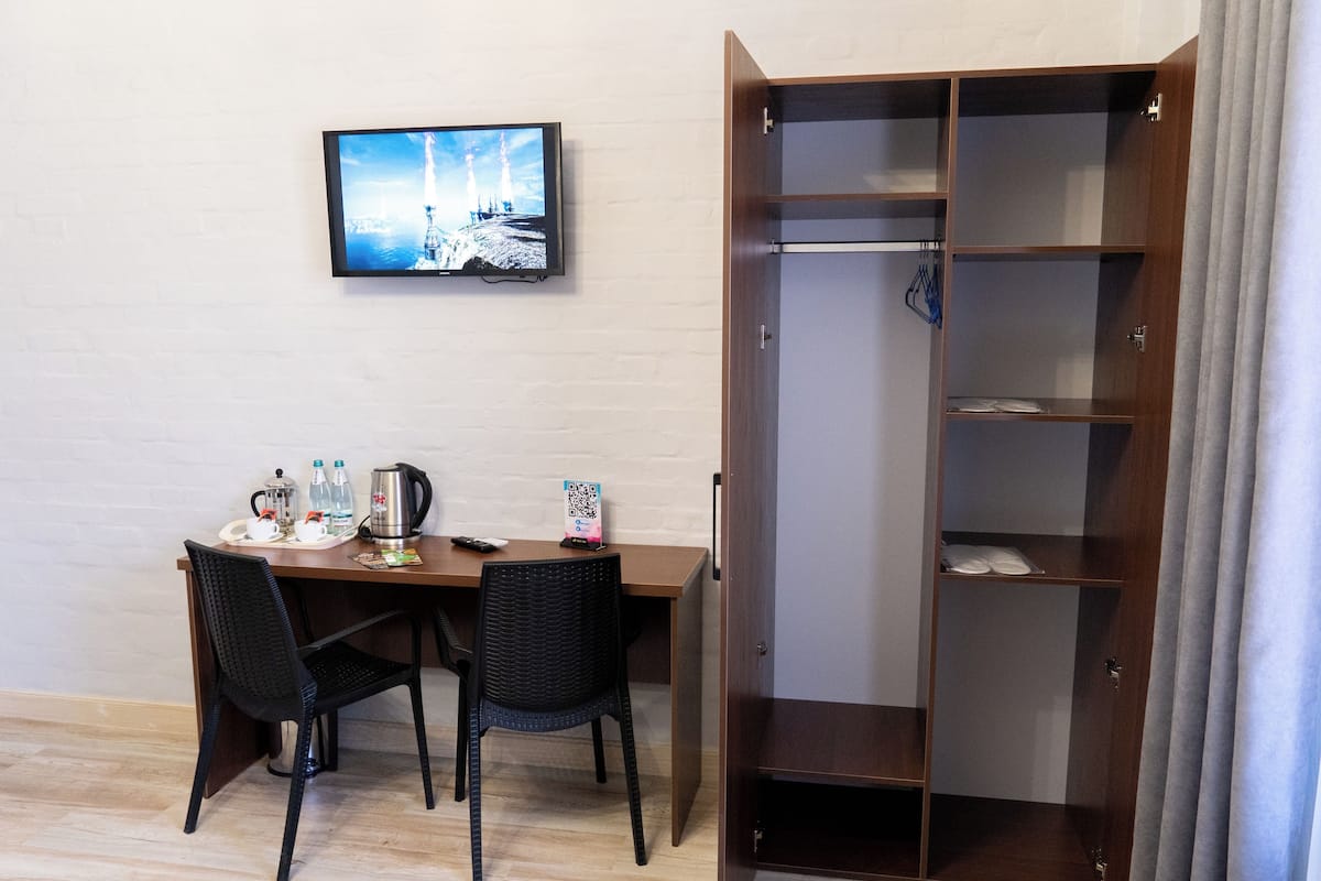 A hotel room with a wooden desk, two chairs, a television mounted on the wall, and a wardrobe with multiple shelves.