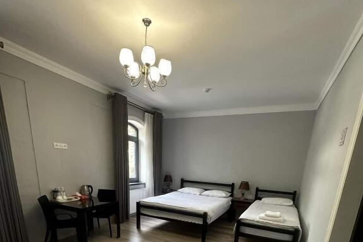 A room with two single beds, a dining table with chairs, a chandelier, and a window with curtains.