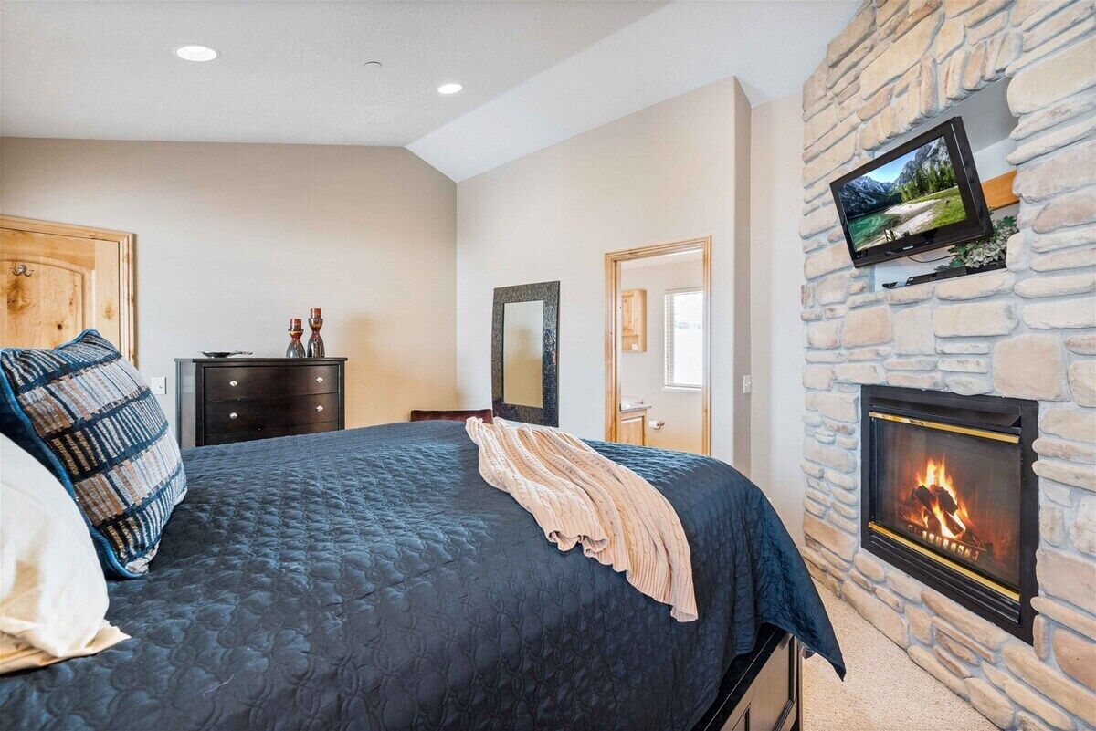 A bedroom with a large bed, a dresser, a mirror, a television mounted on a stone wall, and a fireplace.