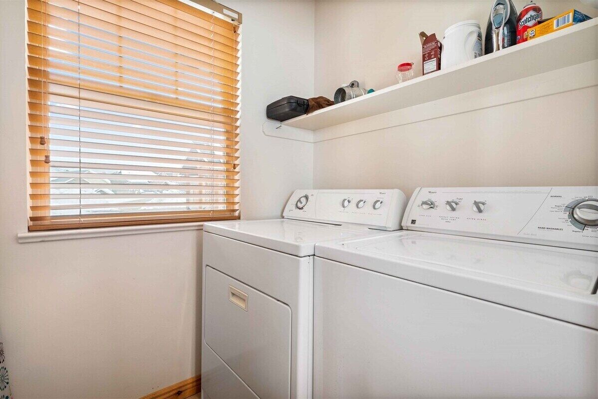 A laundry room with two front-loading washing machines, a shelf with cleaning supplies, and a window with blinds.