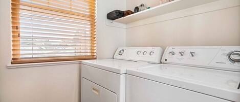 A laundry room with two front-loading washing machines, a shelf with cleaning supplies, and a window with blinds.