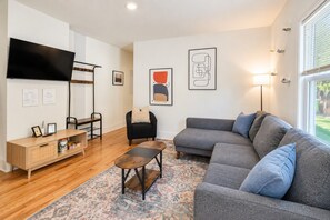 Una sala moderna con un sofá seccional gris, una mesa de centro de madera, un sillón negro, un televisor montado en la pared y una alfombra estampada.