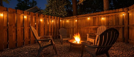 An outdoor seating area with wooden chairs and a fire pit, surrounded by a wooden fence and string lights.