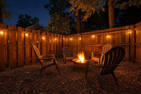 An outdoor seating area with wooden chairs and a fire pit, surrounded by a wooden fence and string lights.