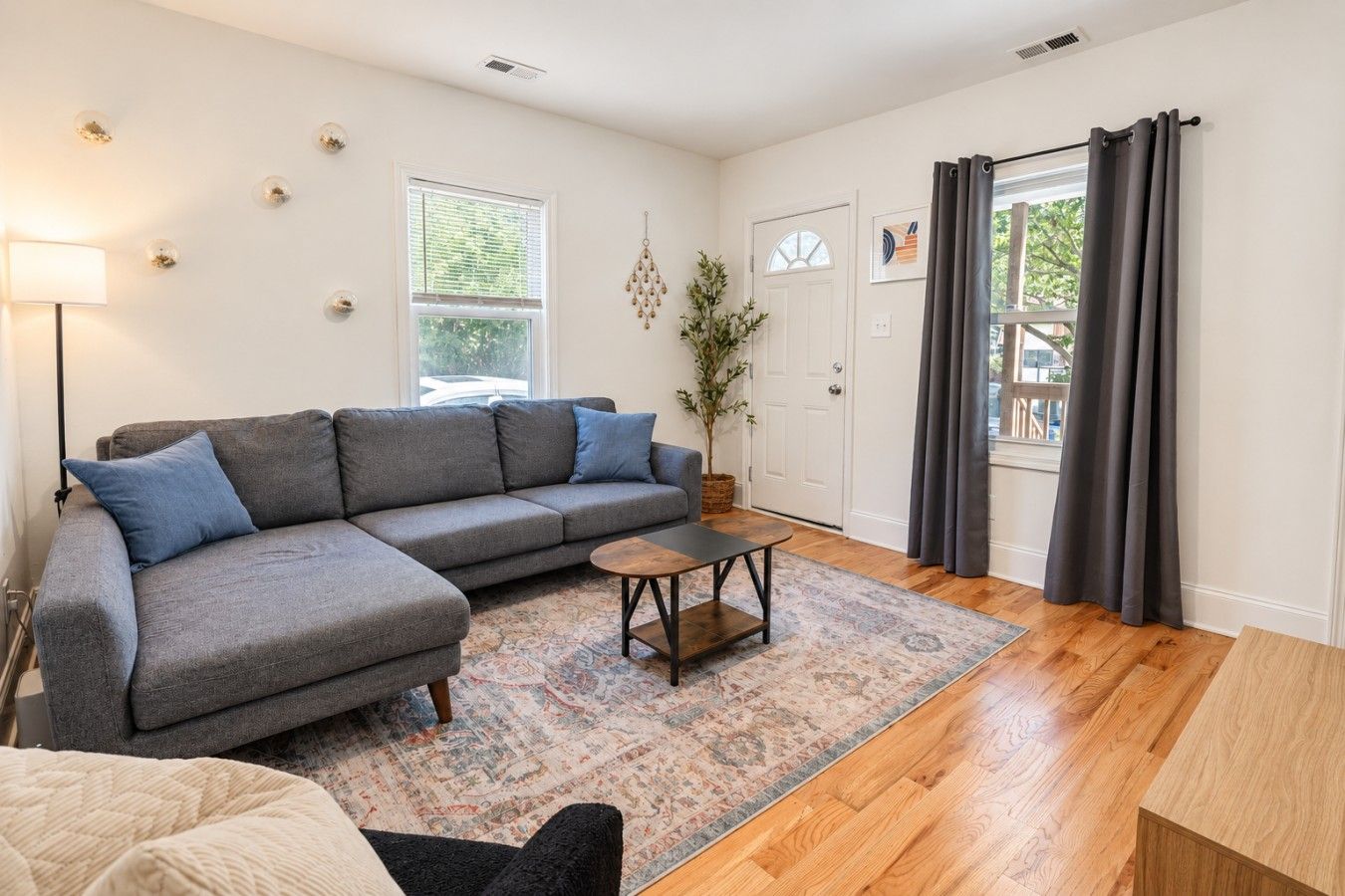 A living room with a grey sectional sofa, a small round coffee table, a patterned rug, wooden flooring, a floor lamp, and a door with curtains.