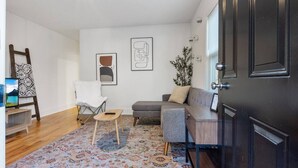 Un salon avec un canapé gris, un fauteuil blanc, une petite table en bois, un tapis à motifs et une porte sombre.