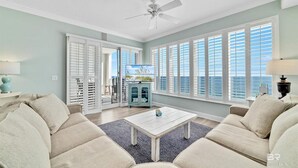 A spacious living room with a white ceiling fan, large windows with shutters, a light blue wall, and a view of the ocean. - Regency Isle 1102 3 3 2 Covered Parking Spots Penthouse (Orange Beach)