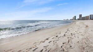 Una spiaggia sabbiosa con impronte che conducono all'acqua, un cielo sereno e sullo sfondo la linea dell'orizzonte di una città.