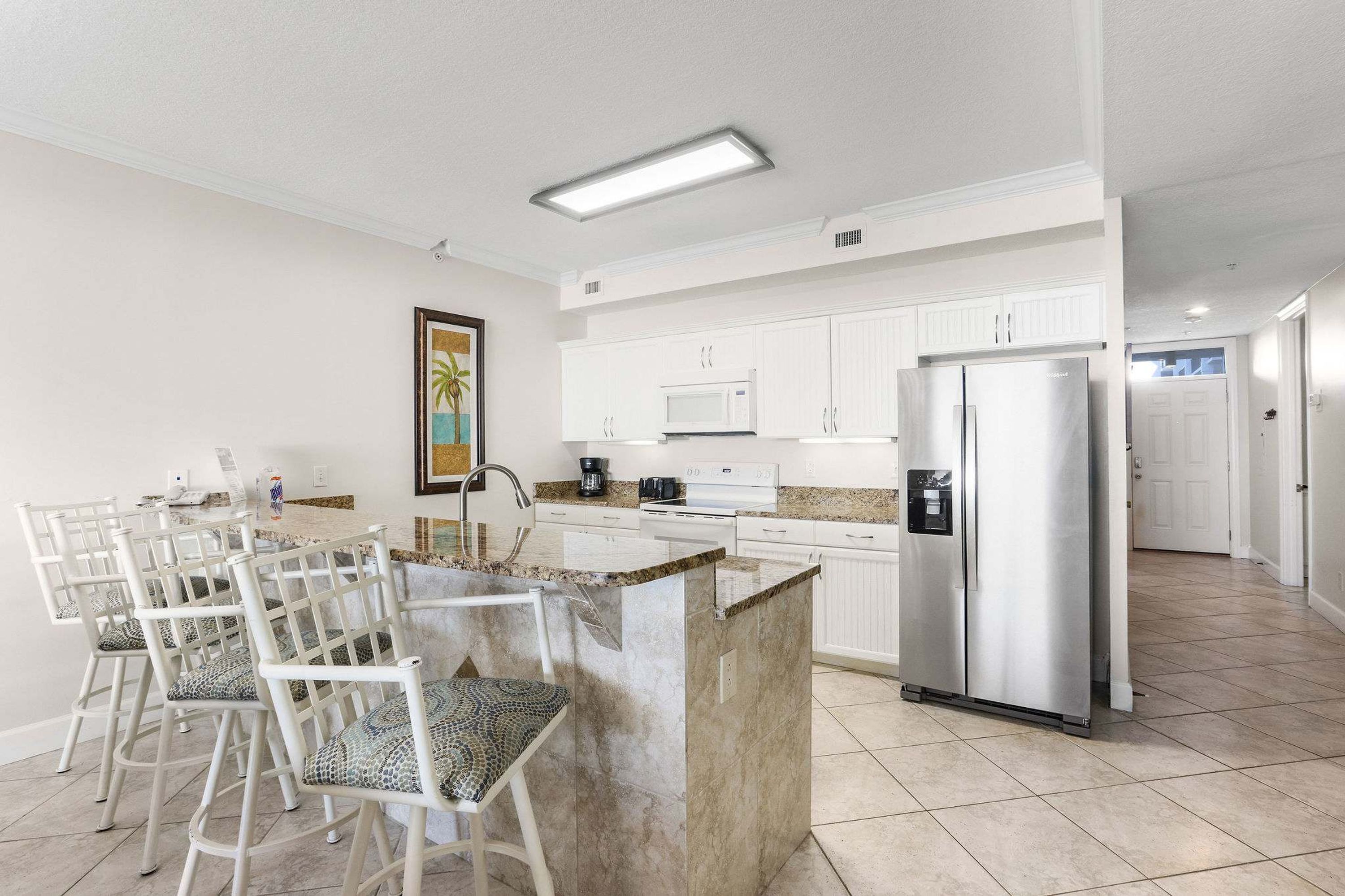 A modern kitchen with white cabinetry, a stainless steel refrigerator, and a granite countertop island with bar stools.