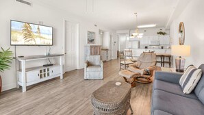 A spacious living room with a television, seating area, and a kitchen in the background. - Grand Pointe 512 Gulf-front Stunning Views Top-notch Amenities (Orange Beach)