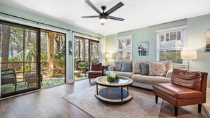 A spacious living room with a large sliding glass door leading to a garden, a ceiling fan, a sofa set, and a coffee table.