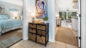 A bedroom with a bed, bedside table, lamp, and a painting. There is a hallway leading to a kitchen area.