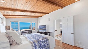 A bedroom with a large bed, a desk, a television, and a view of the sea through a window. - Luxurious Seaside Escape-85 Steps to the Beach-Panoramic Ocean Views (Laguna Beach)