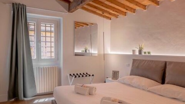 A bedroom with a large bed, a window with curtains, a wooden ceiling, and a nightstand with a lamp.