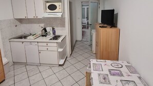 Et kompakt køkken med hvide skabe, en mikrobølgeovn og en kaffemaskine. Der er et lille spiseområde med et bord dækket af en dug, og med billeder i rammer.
