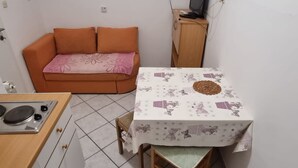 A compact living space with a kitchenette, a dining table with a floral tablecloth, an orange sofa with a pink cushion, and a wooden cabinet with a television on top. - Apartments Pansion Franka 3* - Apartment 10 (Brist)