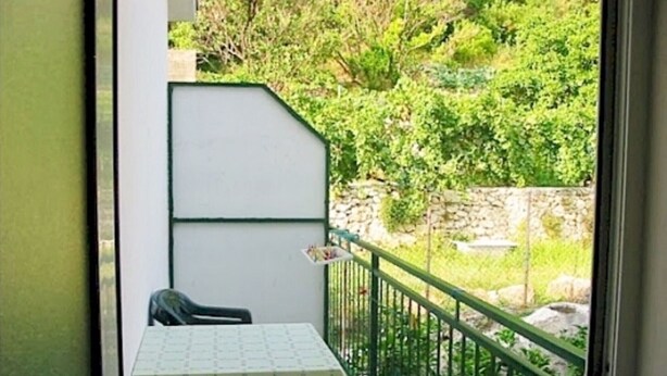 A balcony with a table covered by a patterned tablecloth, a chair, and a view of greenery and a stone wall. - Apartments Pansion Franka 3* - Apartment 10 (Brist)
