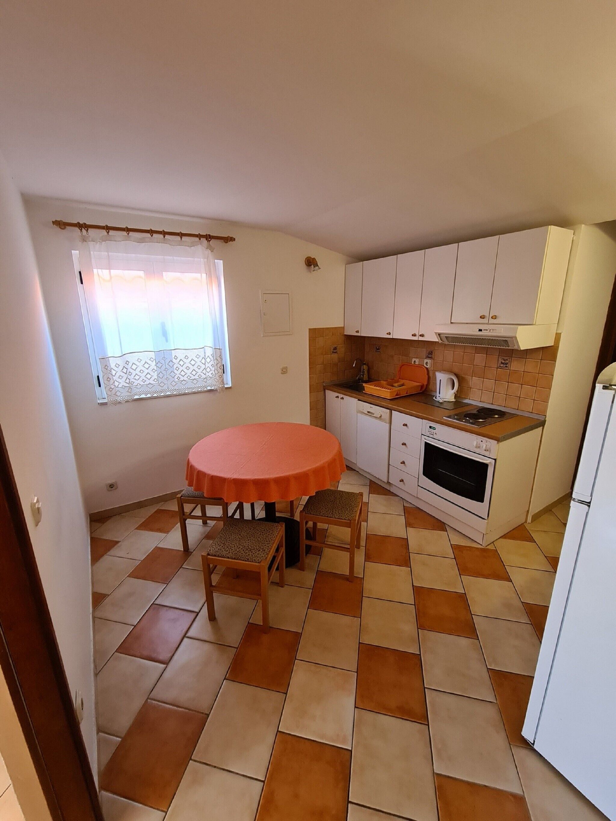 Eine kleine Küche mit einem runden Tisch, zwei Stühlen, einem weißen Kühlschrank, einem Herd und einem Fenster mit Vorhang.