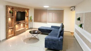 A modern living room with a sectional sofa, a flat-screen TV mounted on a stone wall, and a large window with blinds. - Spacious 3-bedroom apartment in excellent Abuja with AC, WiFi, fitness room (Abuja)